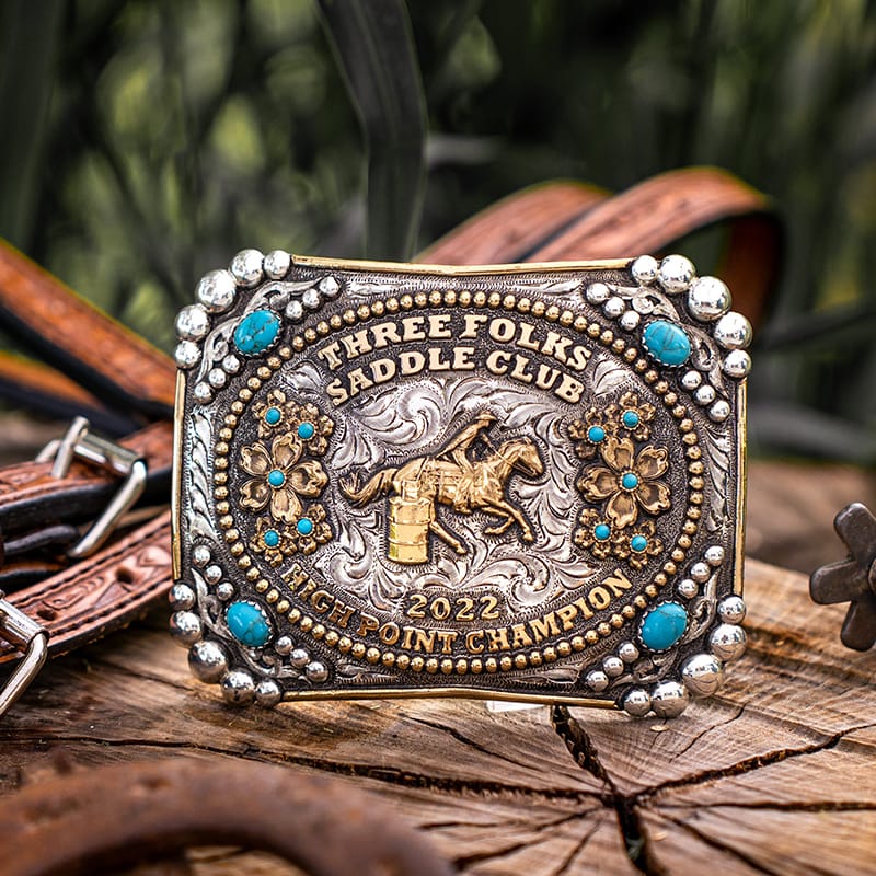 A custom rodeo belt buckle with a barrel racing figure in the center and turquoise stones